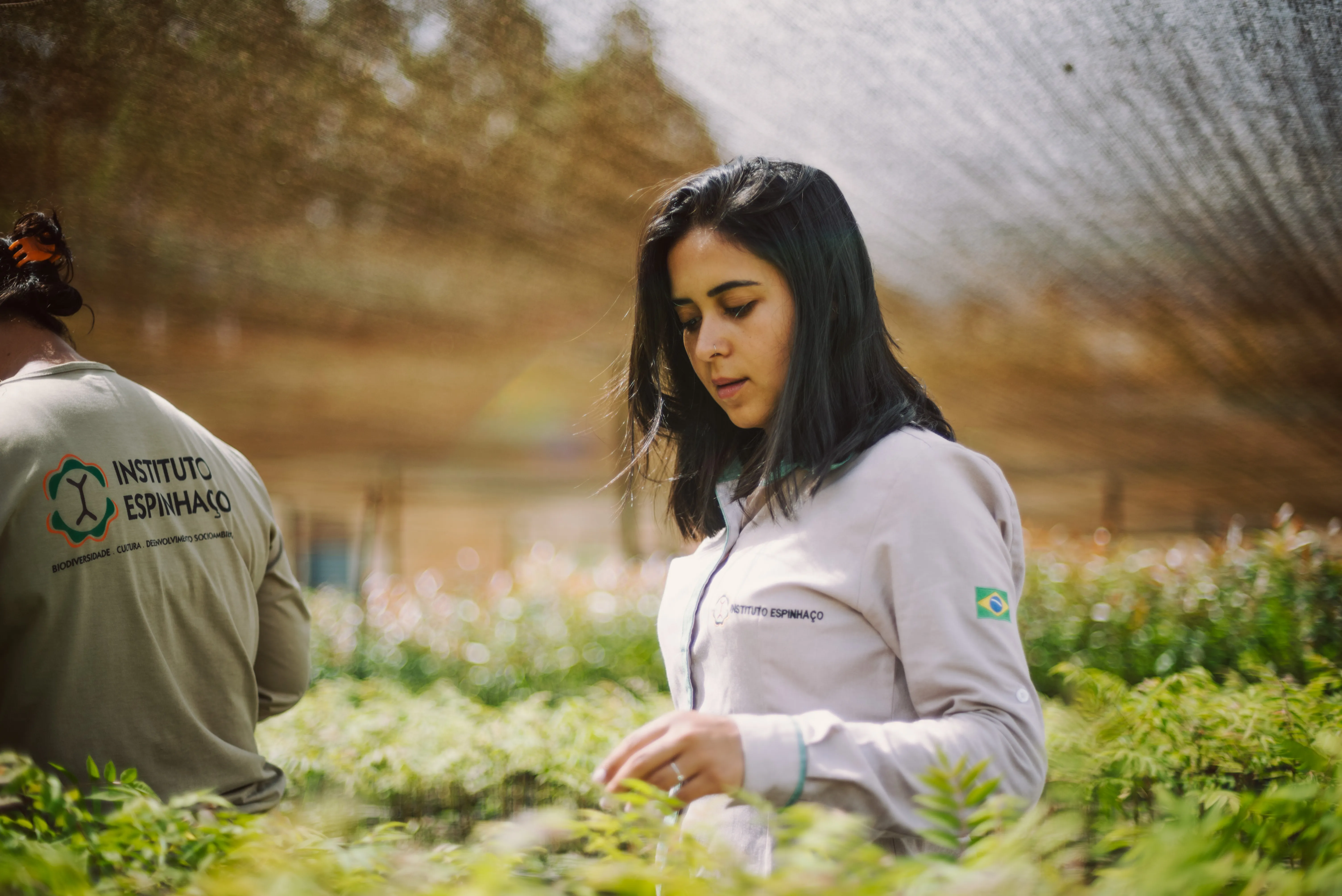 Eine junge weiblich gelesene Person
arbeitet konzentriert in einer Baumschule oder einem Gewächshaus unter einem Schattiernetz. Sie trägt eine helle Arbeitsjacke
mit dem Logo 'Instituto Espinhaço' und einer kleinen Brasilien-Flagge auf dem Ärmel.