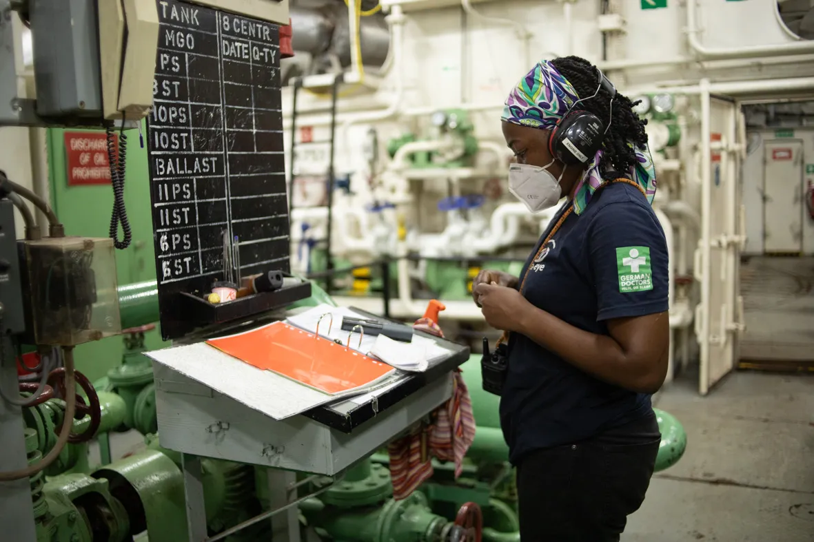 Eine Technikerin
mit Atemschutzmaske, Gehörschutz und buntem Kopftuch steht im Maschinenraum des Schiffes SEA-EYE 4. Sie blickt auf
einen Arbeitstisch mit Ordnern vor einer großen Tafel, auf der Betriebsdaten von Tanks notiert sind.