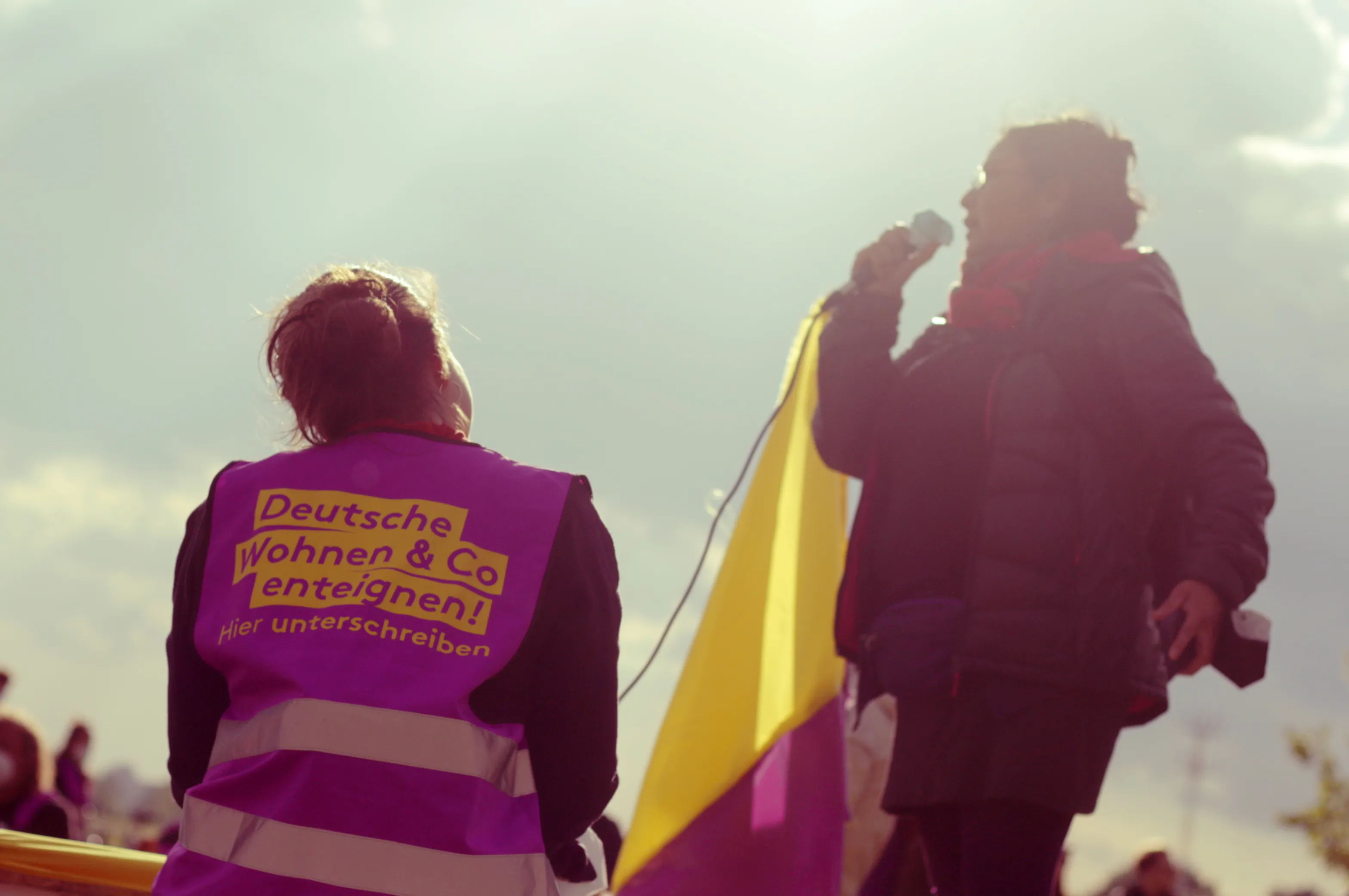 Redebeitrag: Rückansicht einer Person bei einer Kundgebung,
die eine violette Weste mit der gelben Aufschrift 'Deutsche Wohnen & Co enteignen! Hier unterschreiben' trägt.
Im Hintergrund spricht eine weitere Person in ein Mikrofon vor einer gelb-violetten Fahne.